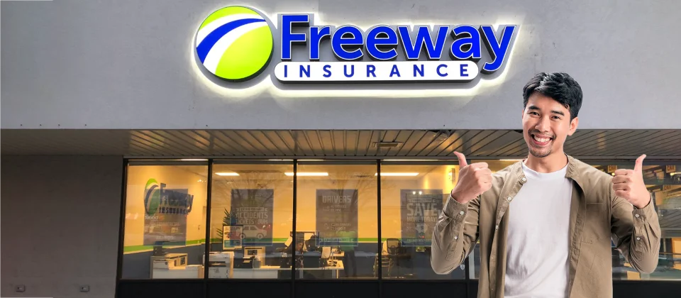 A smiling young Hispanic man stands in front of a Freeway Insurance office showing the thumbs up signal with his hands.